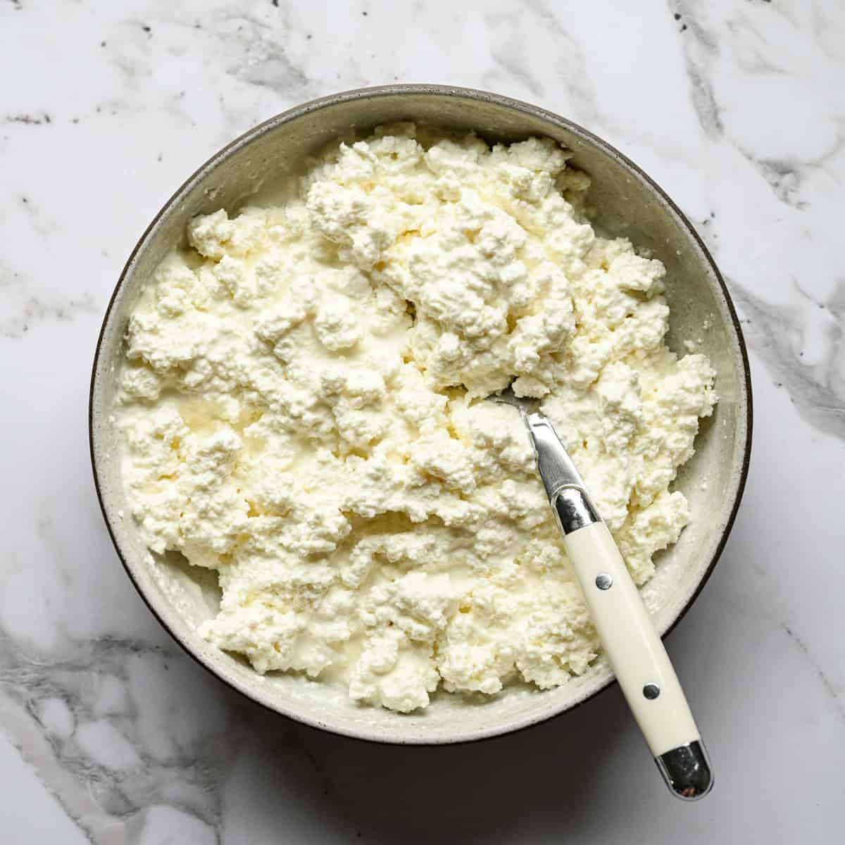 Overhead view of creamy homemade cottage cheese in a bowl with a spoon stirring in heavy cream