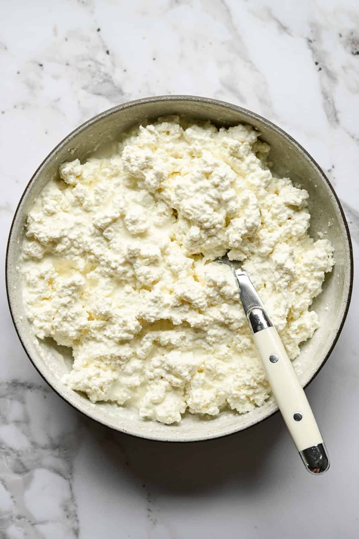 Overhead view of a bowl of homemade cottage cheese with a spoon on a marble counter