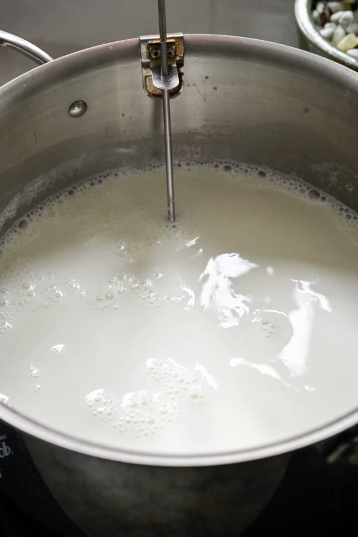 Whole milk heating in a large stainless steel pot with a clip-on thermometer