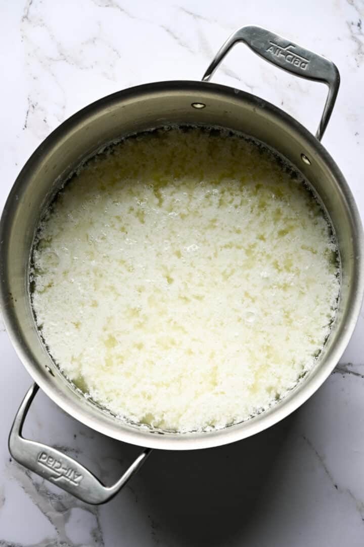 Overhead view of cottage cheese curds separated from whey in an All-Clad pot on marble