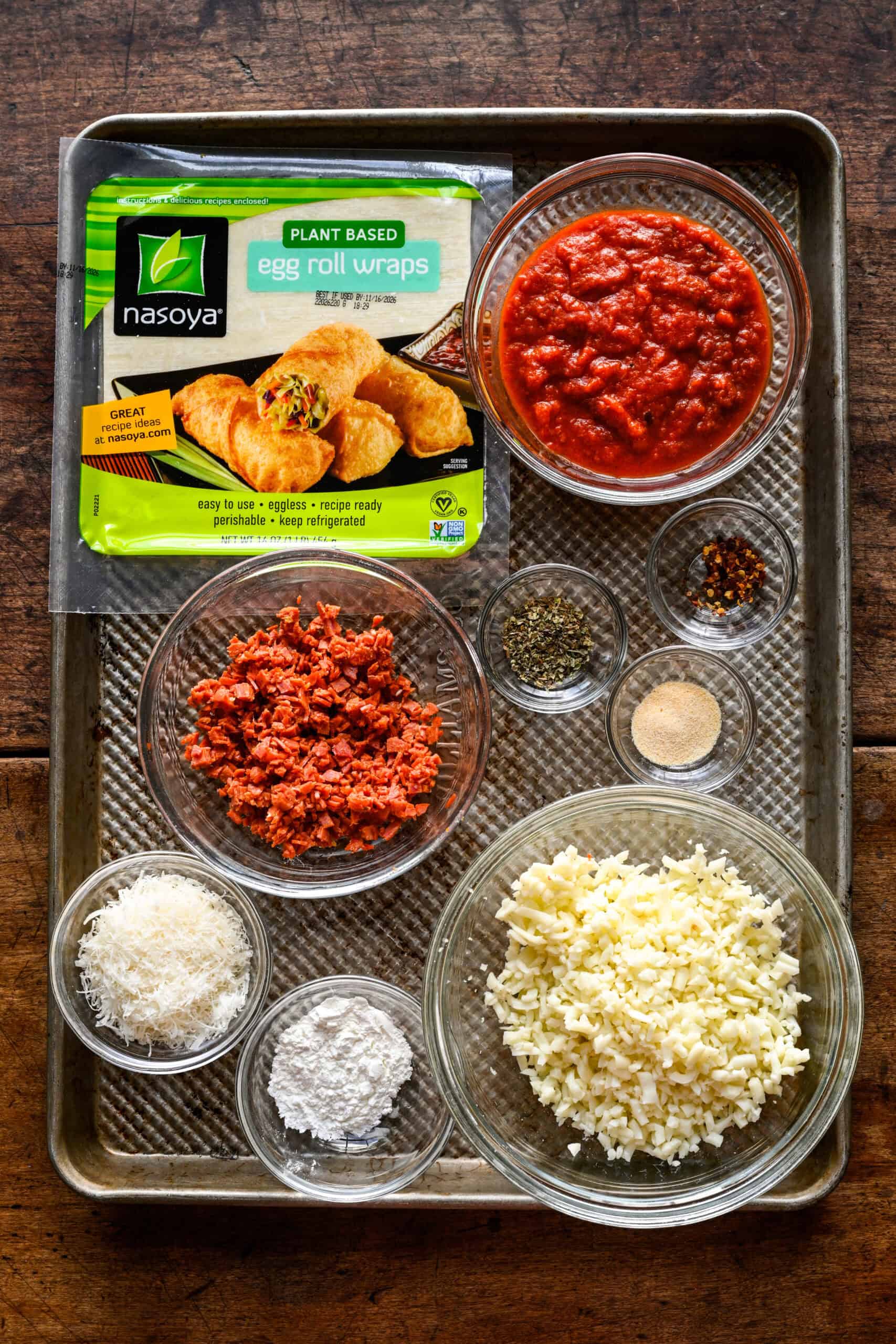 Ingredients for homemade pizza rolls on a sheet pan: a package of egg roll wrappers and bowls of pizza sauce, chopped pepperoni, chopped mozzarella, grated parmesan, cornstarch, Italian seasoning, garlic powder and red pepper flakes.