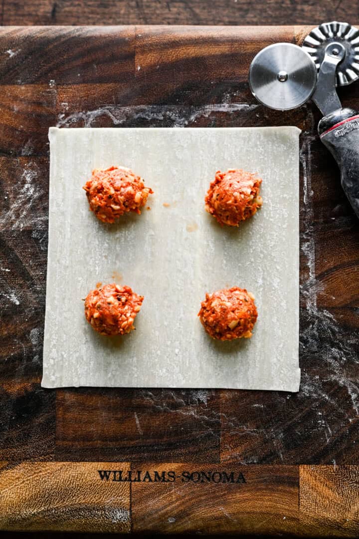 An egg roll wrapper on a wooden cutting board with four evenly spaced mounds of pizza roll filling, one in each quadrant.