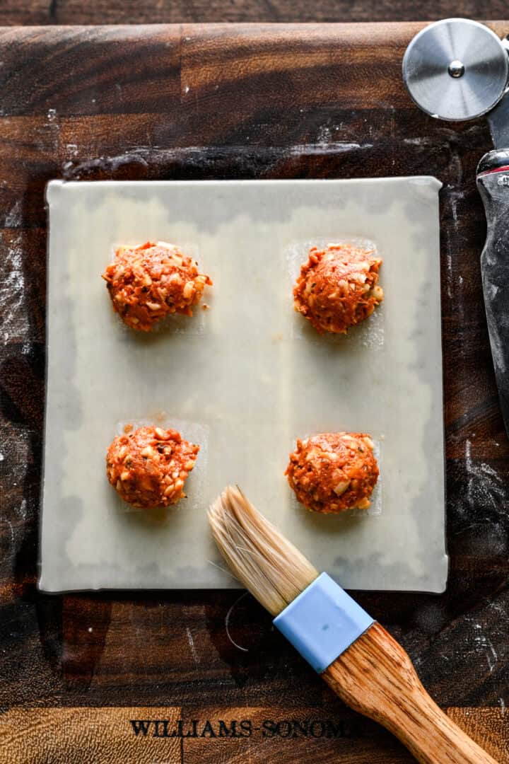 An egg roll wrapper brushed with water around four mounds of pizza roll filling, with a pastry brush resting on the corner.