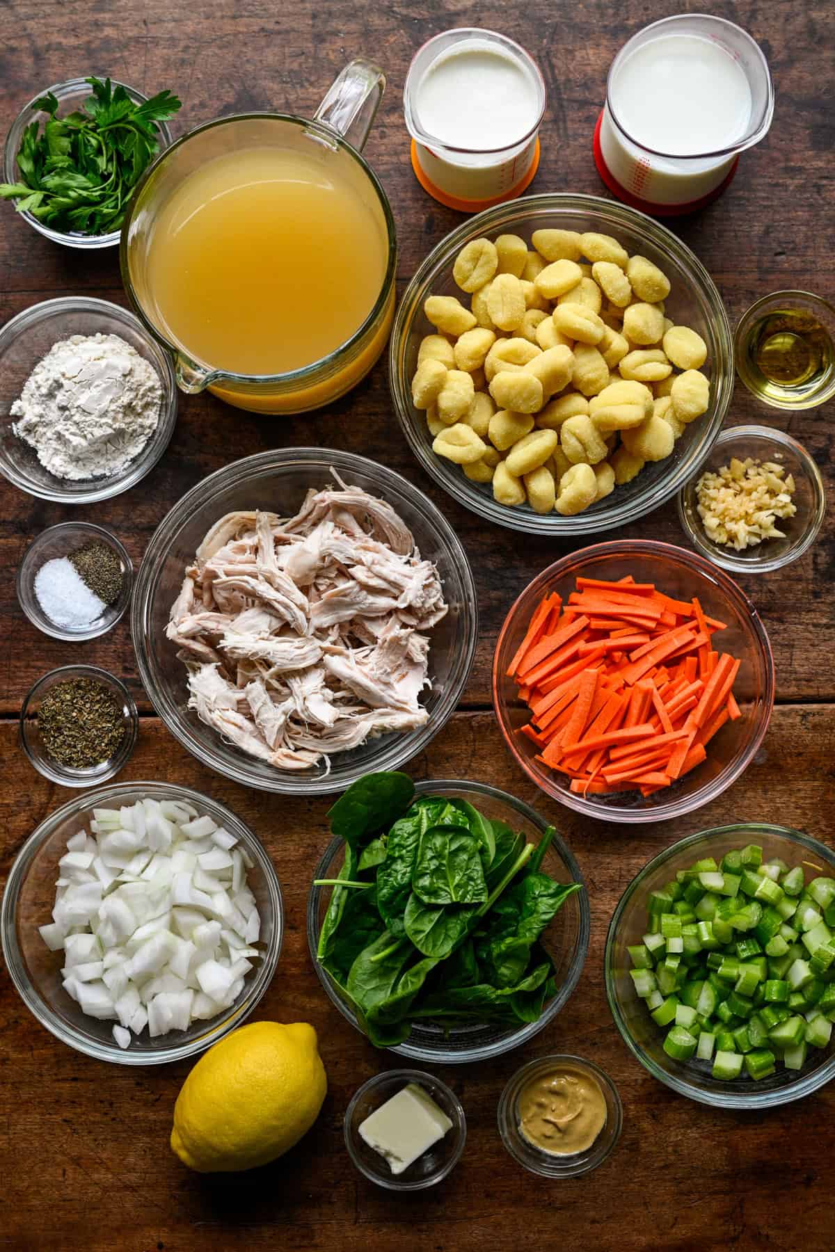 Overhead view of ingredients for chicken gnocchi soup on a wood table: chicken broth, potato gnocchi, shredded chicken, matchstick carrots, spinach, diced onion and celery, flour, milk, cream, parsley, lemon, butter and Dijon mustard in glass bowls