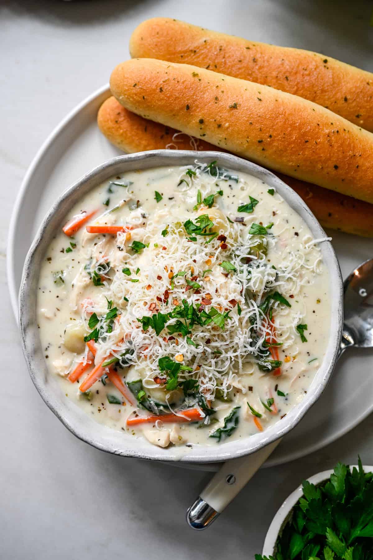 Overhead view of a bowl of chicken gnocchi soup topped with parmesan, parsley and red pepper flakes, with breadsticks and a spoon on the plate