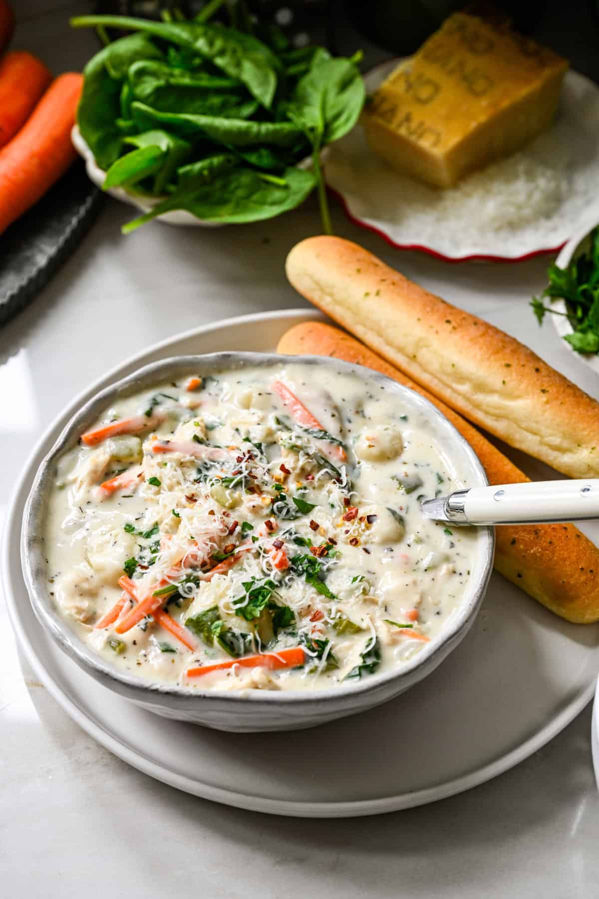 45 degree view of a bowl of chicken gnocchi soup showing shredded chicken, gnocchi, carrots and spinach, with breadsticks and a white spoon