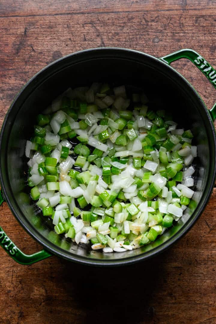 Overhead view of diced onion and celery sautéing in butter in a green Staub Dutch oven