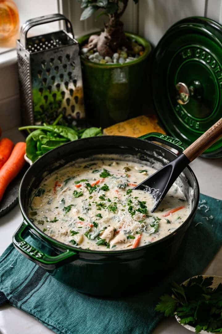 45 degree view of chicken gnocchi soup in a green Staub Dutch oven with a ladle, topped with parsley and grated parmesan, a box grater and spinach behind