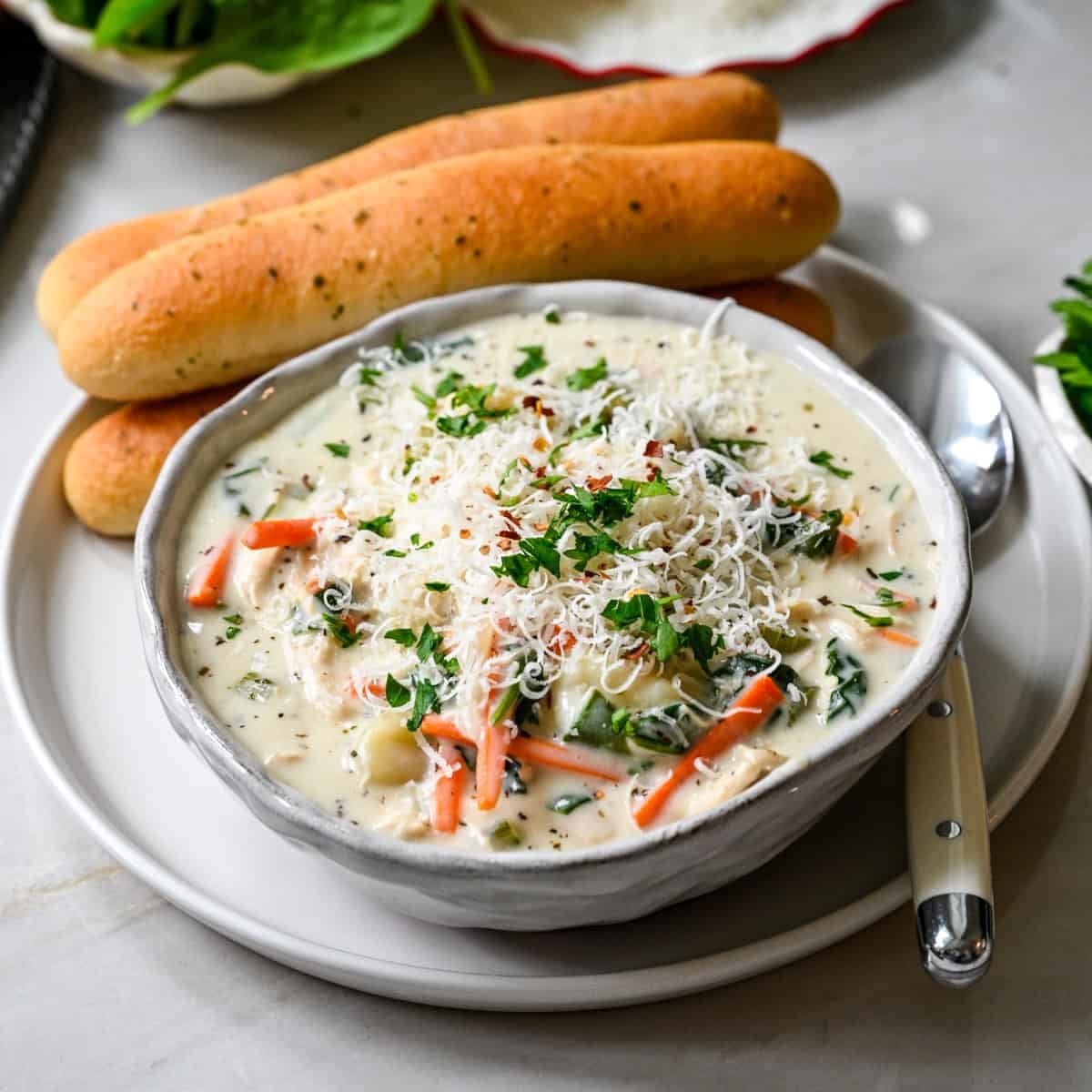 Close up view of creamy chicken gnocchi soup in a gray bowl with shredded parmesan, parsley and red pepper flakes, breadsticks and a spoon beside it