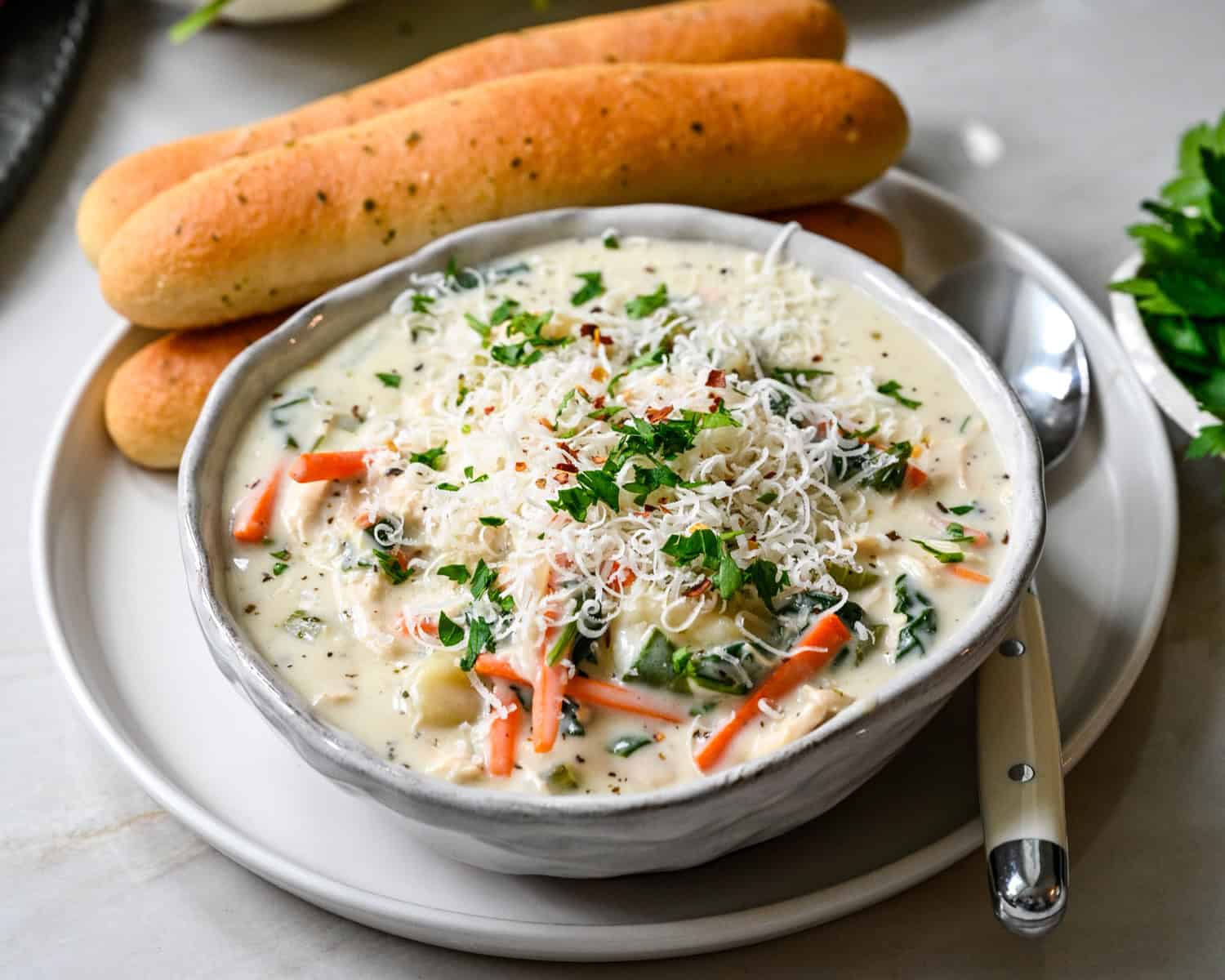 Front view of a bowl of copycat Olive Garden chicken gnocchi soup with carrots, spinach and parmesan, served with breadsticks on a white plate