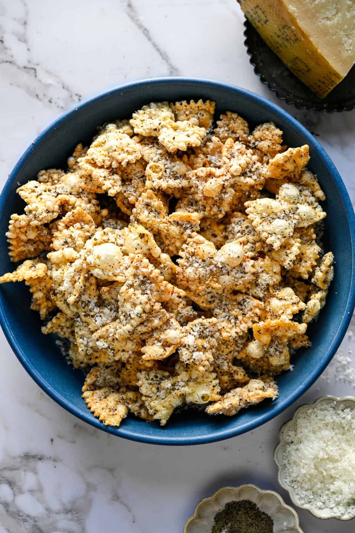 Overhead view of puffed pasta in a blue bowl beside a wedge of parmesan and a dish of pepper.