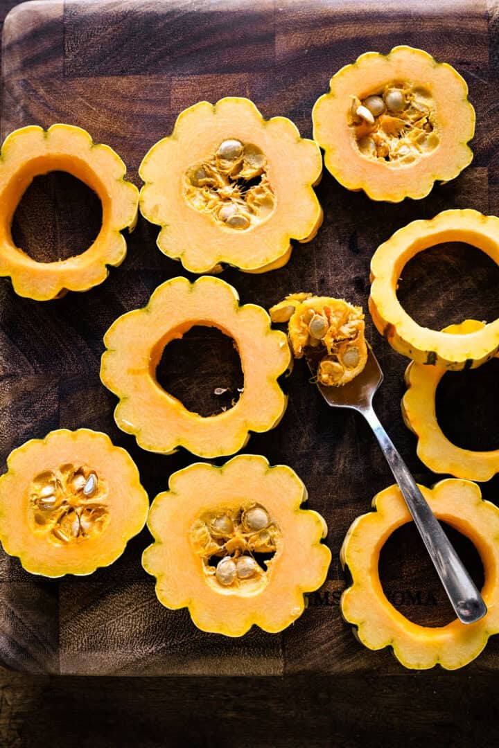 Overhead view of delicata squash rounds on a wooden board, the seeds scooped out with a spoon.