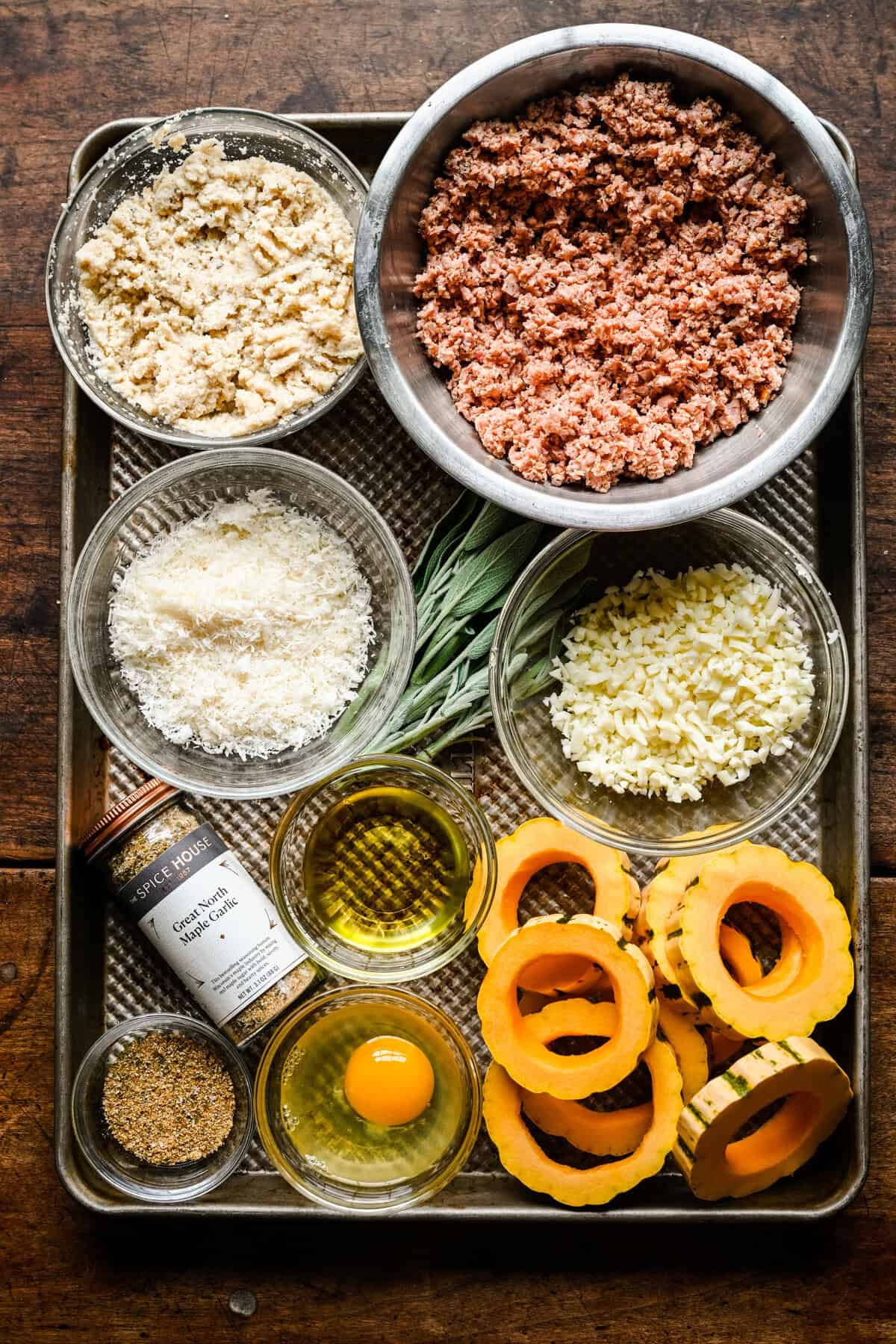 Overhead view of ingredients on a sheet pan: ground sausage, breadcrumbs, parmesan, mozzarella, fresh sage, olive oil, an egg, Great North Maple Garlic seasoning and delicata squash rings.