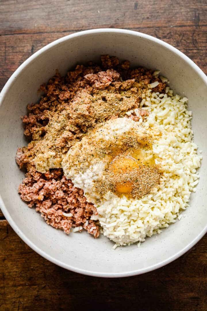 Overhead view of ground sausage, mozzarella, breadcrumbs, an egg and seasoning in a white bowl before mixing.