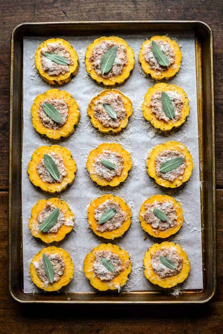 Overhead view of squash rings on a sheet pan filled with sausage filling, sprinkled with parmesan and topped with sage leaves before baking.