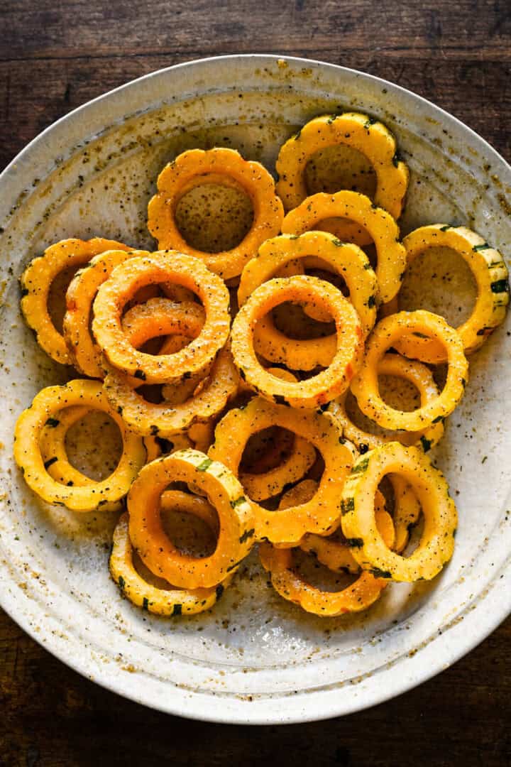 Overhead view of delicata squash rings tossed with oil and seasoning in a white bowl.