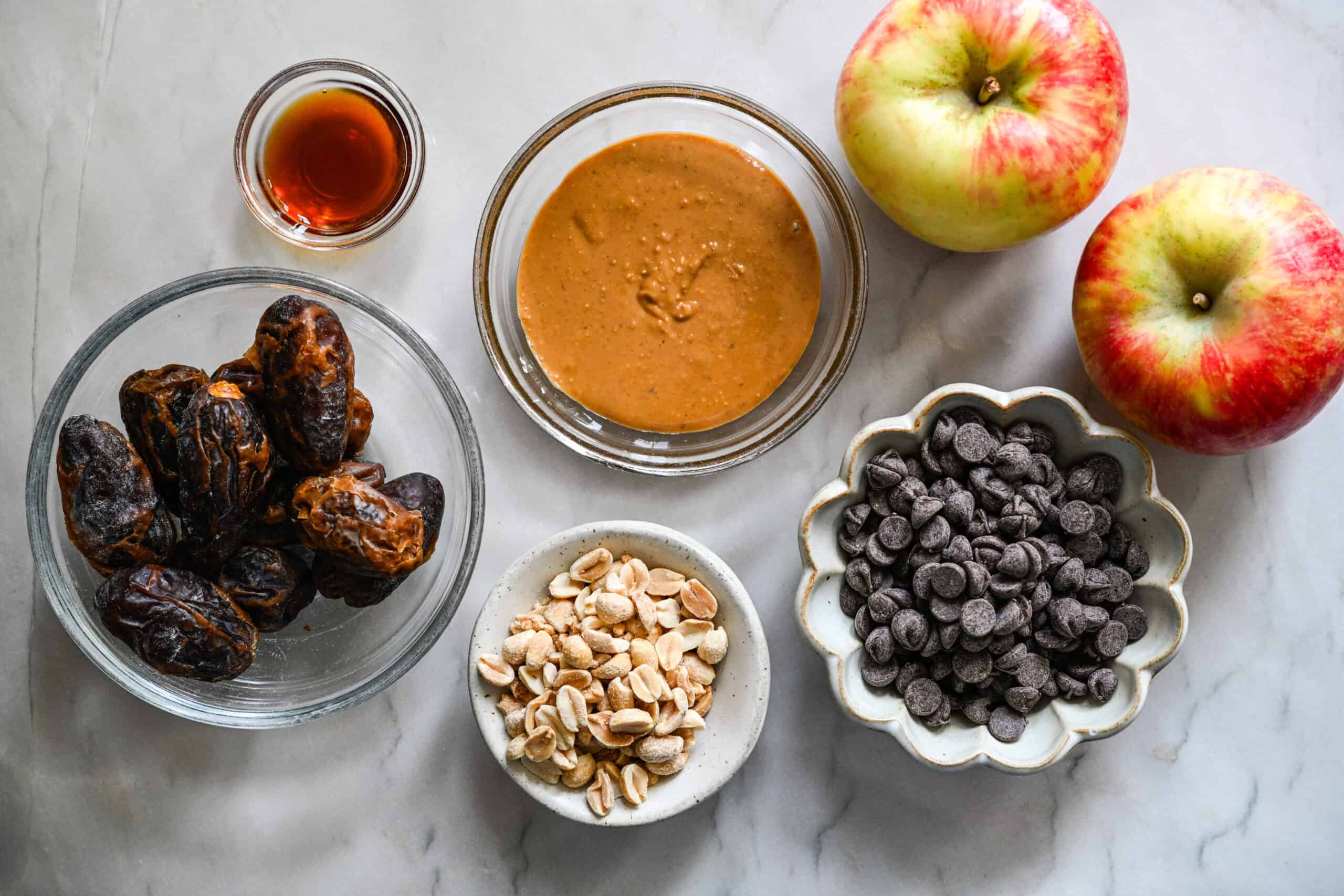 Overhead view of dates, peanut butter, vanilla, peanuts, chocolate chips and two apples on a marble counter.