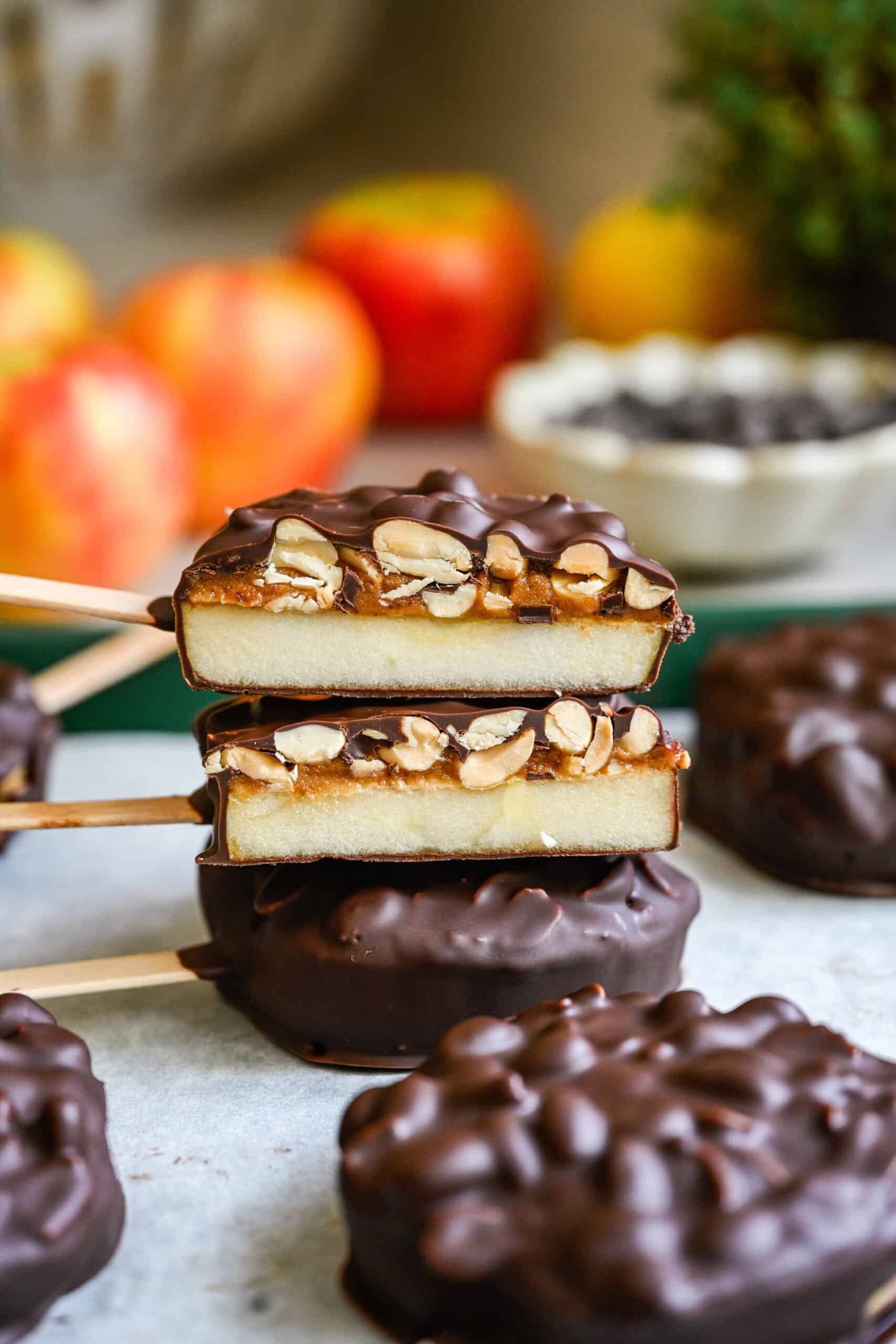 Front view of two halved chocolate covered apple slices stacked to show layers of apple, date spread and peanuts, with apples behind.