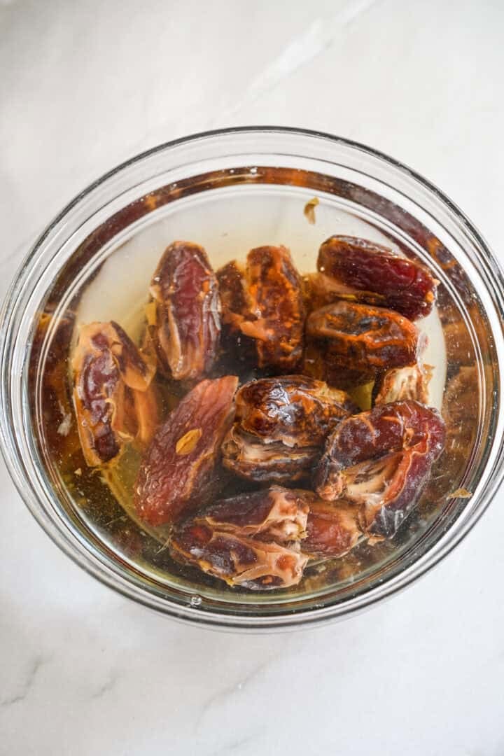 Overhead view of pitted, halved dates soaking in water in a glass bowl.