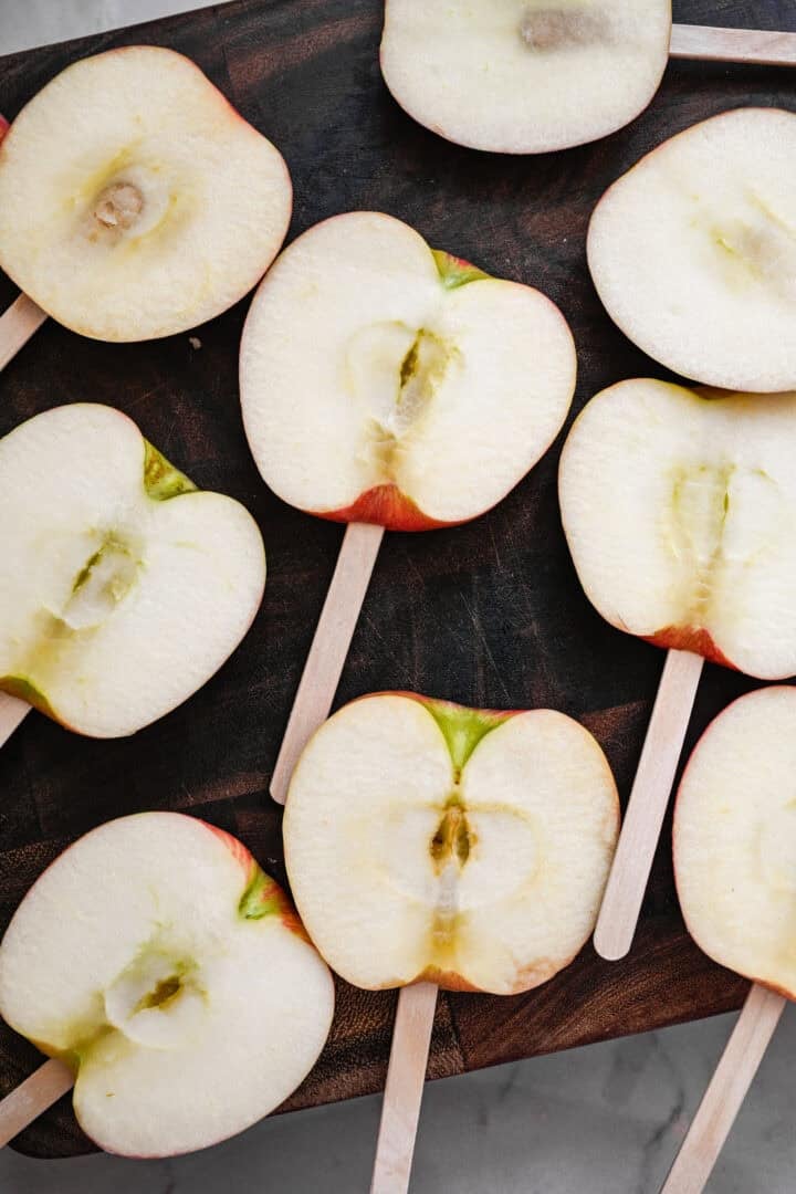 Overhead view of thick apple slices on popsicle sticks on a dark wood cutting board.