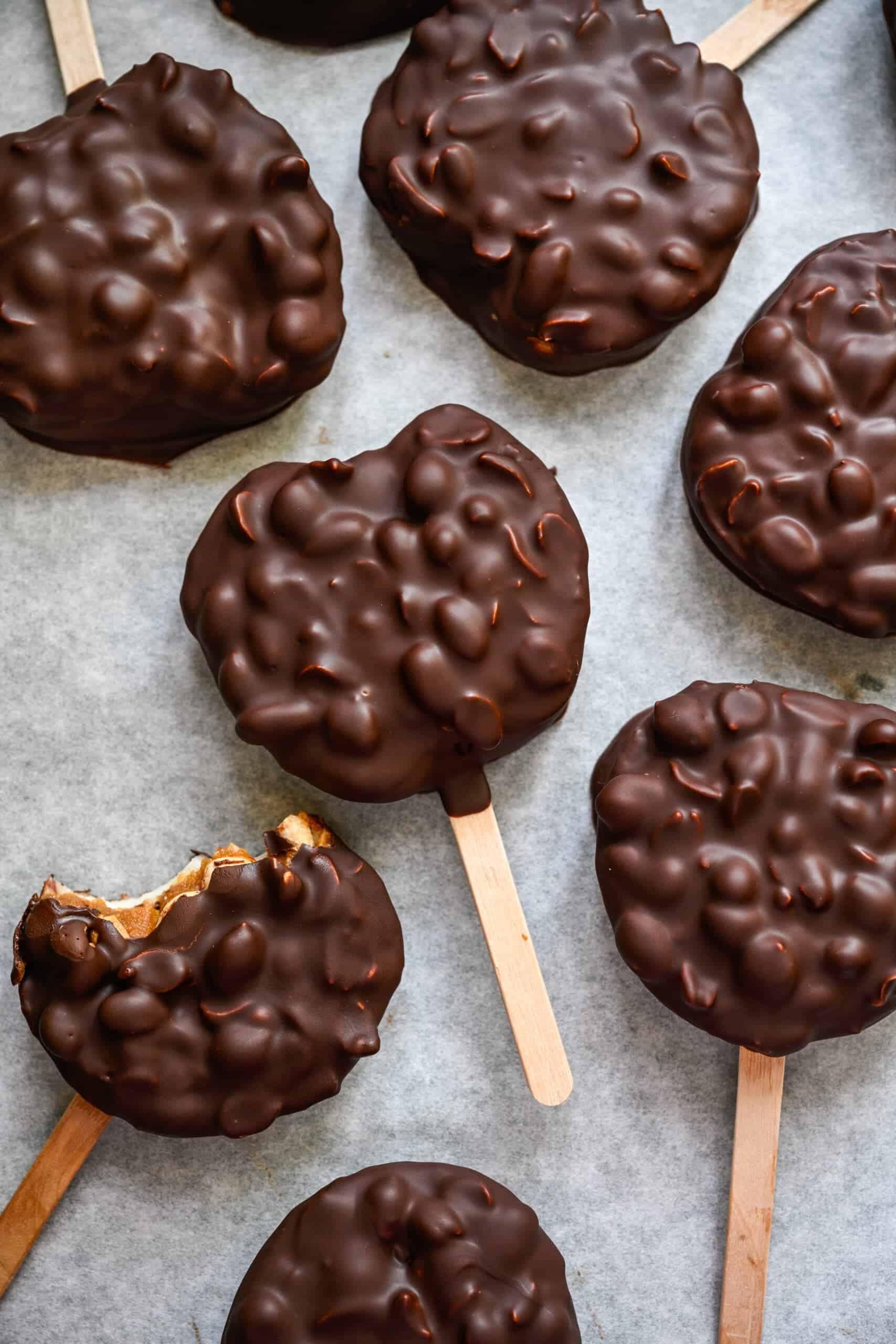 Overhead view of chocolate covered apple slices on popsicle sticks on parchment paper, one with a bite taken out.