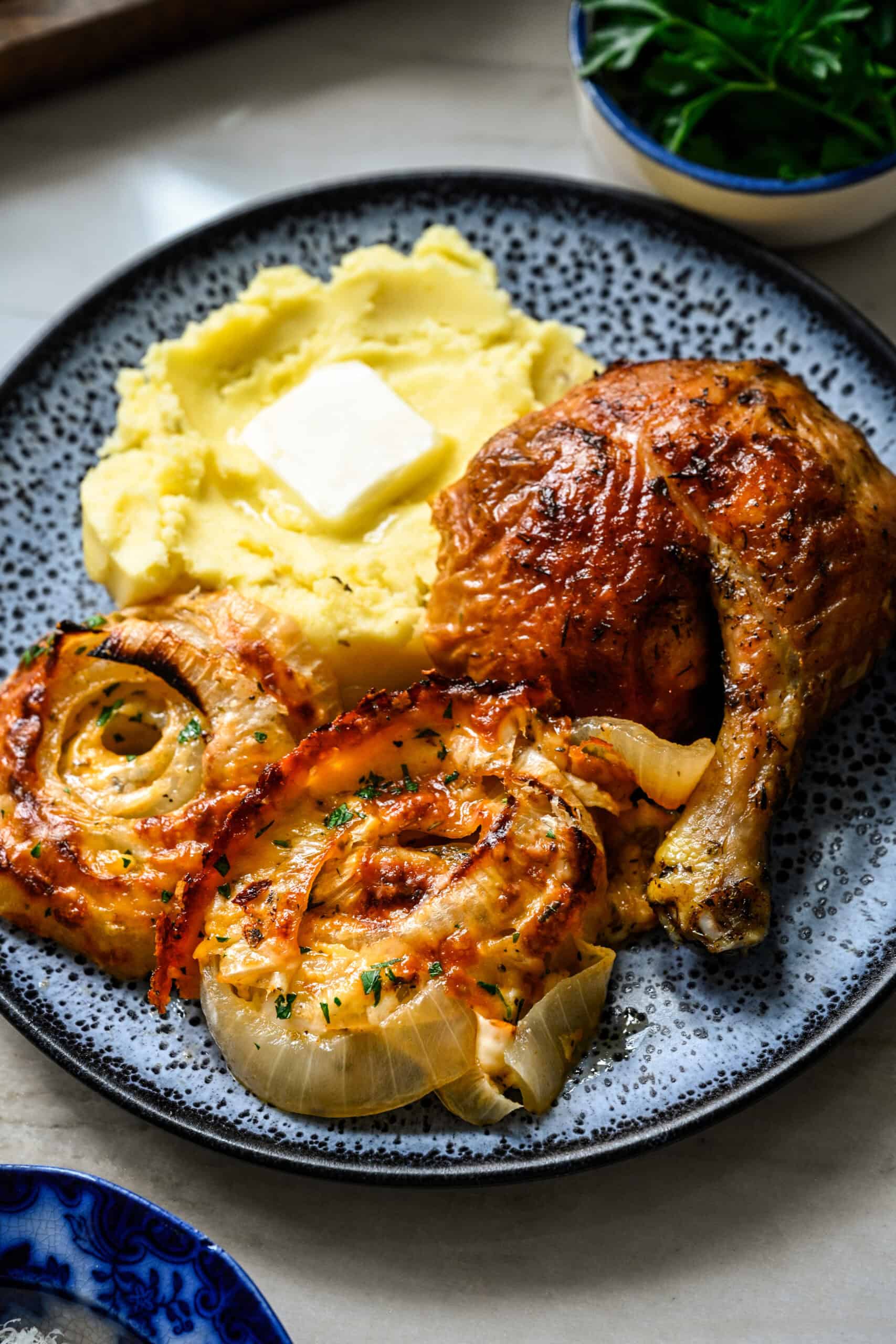 45 degree view of a dinner plate with two scoops of Tennessee onions, mashed potatoes with butter, and a roasted chicken leg quarter.