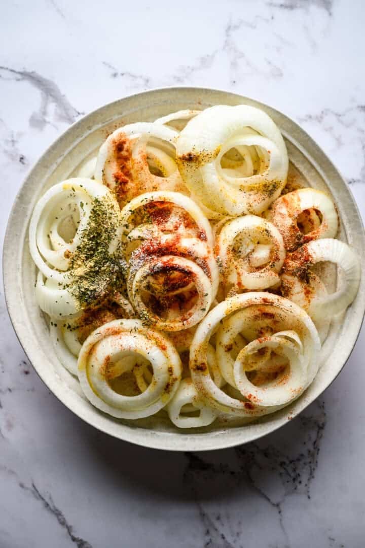 Overhead view of raw sweet onion rings in a wide ceramic bowl, sprinkled with dried herbs, smoked paprika and salt before tossing.