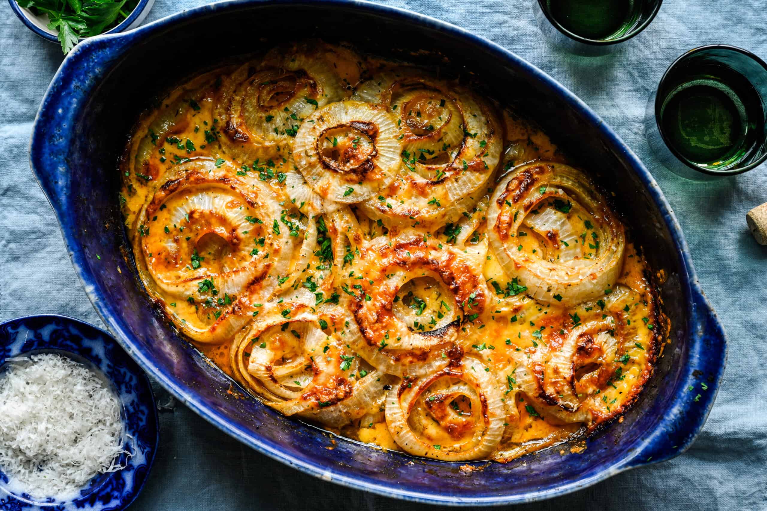 Overhead view of finished Tennessee onions in a blue oval baking dish, garnished with chopped parsley, with grated Parmesan and green glasses alongside on a blue linen.