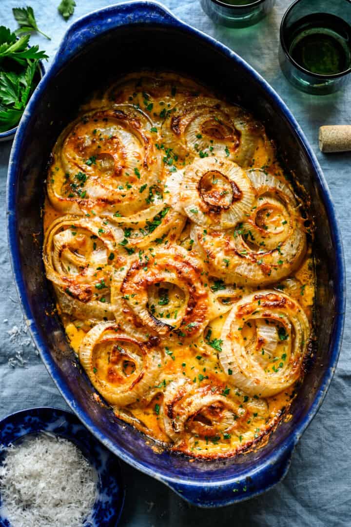 Overhead view of Tennessee onions in a blue oval baking dish with golden brown cheese and chopped parsley, with bowls of fresh parsley and grated Parmesan on the side.